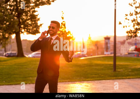 A handsome young businessman walking in a park talking on his phone on a sunny day. Stock Photo