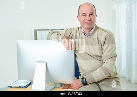 Portrait of cheerful middle-aged businessman sitting on edge of his office desk and leaning on computer screen Stock Photo