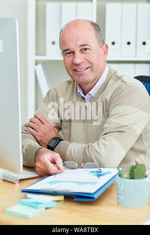 Positive mature financial manager sitting at his office table, taking off glasses and looking at camera Stock Photo