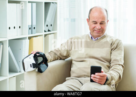 Smiling mature man sitting on sofa and watching tutorial on smartphone before using virtual reality headset for the first time Stock Photo