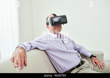 Portrait of smiling mature entrepreneur in lavender shirt resting on sofa in virtual reality glasses Stock Photo