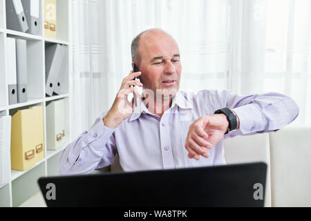 Middle-aged businessman sitting at office desk and checking time on his smartwatch when talking on phone with business partner or colleague Stock Photo