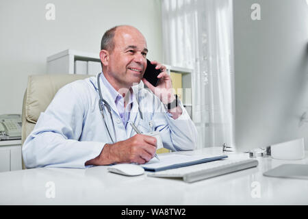 Smiling doctor reading information on computer screen and taking notes when talking on phone with patient Stock Photo