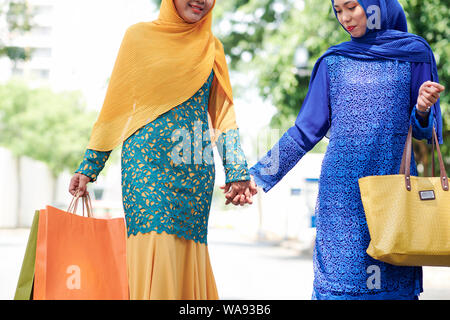 Positive young muslim women in hijab and beautiful dresses holding hands when walking outdoors after shopping in mall Stock Photo