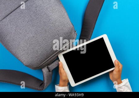 Kid hands with tablet computer and gray backpack over light blue background Stock Photo