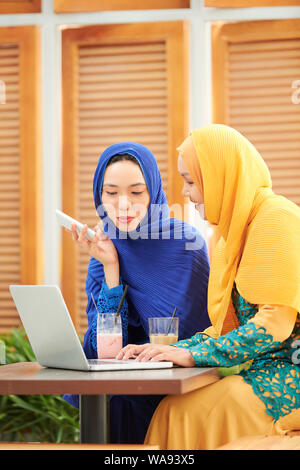 Pretty young muslim woman in hijabs talking and typing on laptop when working on project for university Stock Photo