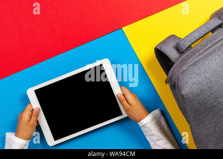 Top view to kid hands with tablet computer and gray backpack on colorful background Stock Photo
