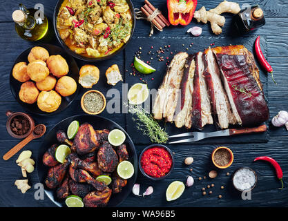 set of caribbean dishes, jerk pork belly, chicken curry, fried dumplings, roasted chicken thighs and drumsticks on plates on a black wooden table with Stock Photo