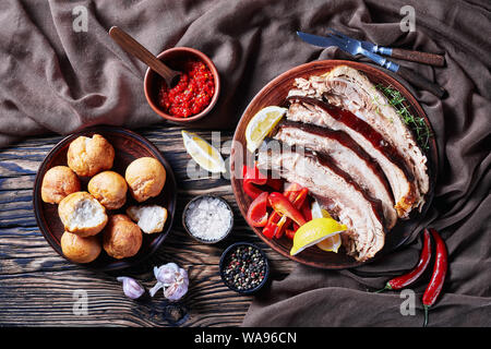 slices of jerky pork belly with lemon wedges and bell pepper on an earthenware plate on a wooden table with brown cloth, horizontal view from above, f Stock Photo