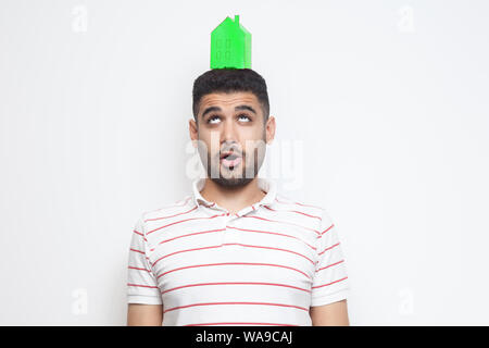 Portrait of thoughtful young man in t-shirt holding green paper small house on head and looking up. Visualize your dreams. Indoor, isolated, studio sh Stock Photo