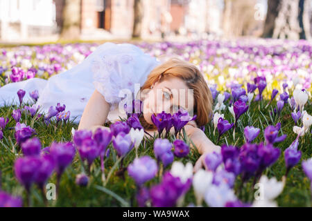 Beautiful young blonde woman with blue eyes wearing in white dress lying on the carpet among the spring flowers crocuses. Spring sunny day. A field of Stock Photo