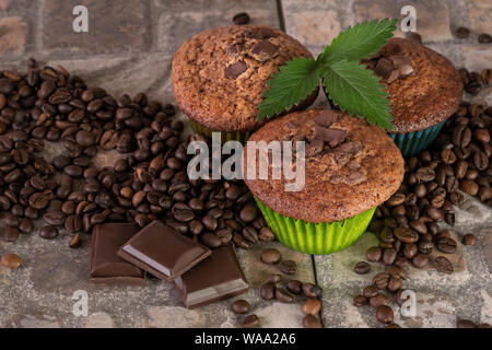 Three muffins with a green sprig and pieces of chocolate surrounded by coffee beans on an earthenware ground. Stock Photo