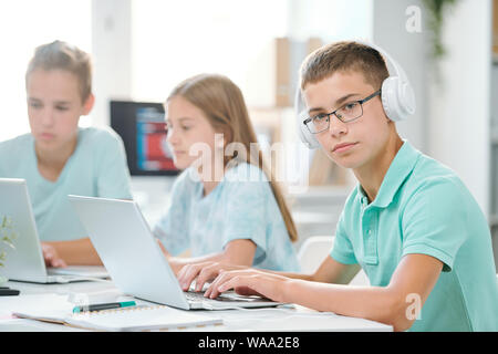 Handsome schoolboy in headphones looking at you while typing on laptop keypad Stock Photo