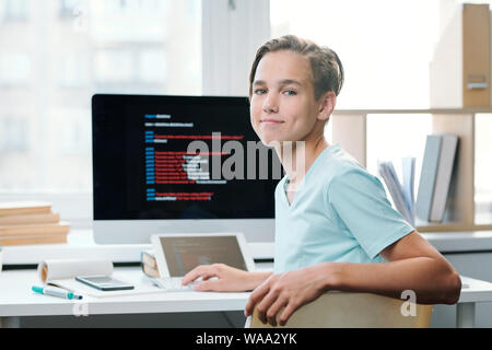 Contemporary boy in casualwear looking at you while sitting by desk Stock Photo