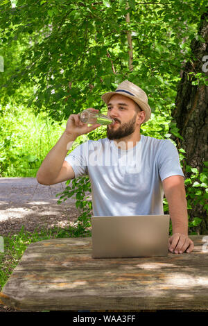 A young attractive man with a beard drinks clear water from a bottle while working at a computer. Sitting in a park at a wooden table Stock Photo