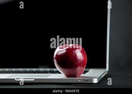 Shiny red apple resting on an open aluminum laptop in selective focus on a black background Stock Photo