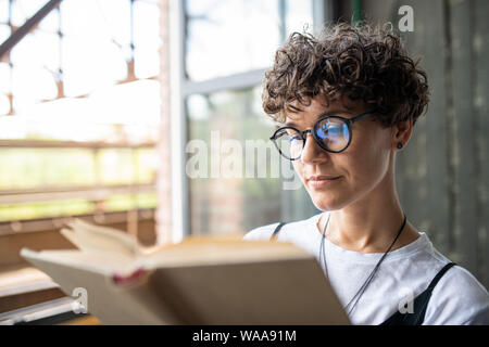 Young serious woman with brown curly hair looking through pages of novel Stock Photo