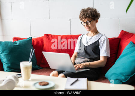 Young busy woman in eyeglasses typing on laptop keypad while looking at display Stock Photo