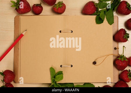 An open notebook of kraft paper with a red pencil are surrounded by strawberries and mint on a light wooden table. Stock Photo