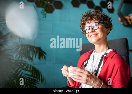 Young cheerful brunette woman with curly hair holding cup of tea or coffee Stock Photo