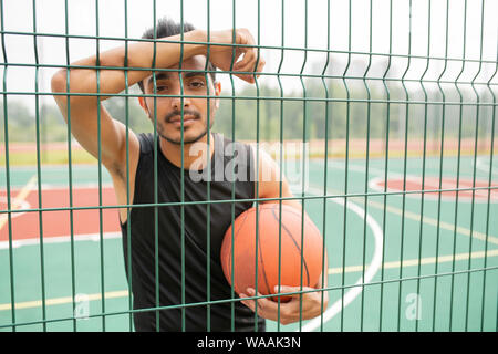 Serious male basketball player with ball standing against net Stock Photo