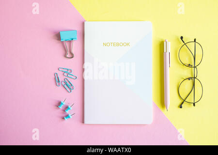 Several pins, clip, notebook, pen and eyeglasses on pink and yellow background Stock Photo