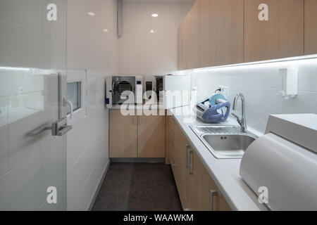 Small room with white walls and a dark gray floor in a dental clinic. There are beige lockers, light tabletop with a sink, autoclave sterilizers, dent Stock Photo