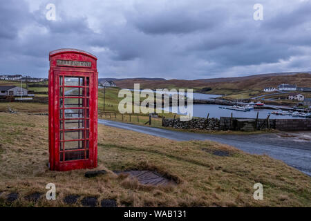 A bright red telephone box stands alone near a road in a rural setting in Shetland, Scotland