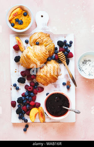 Freshly baked butter croissants with jam, fresh berries and honey on vintage rustic chopping board over pink wooden background. Top view, blank space Stock Photo