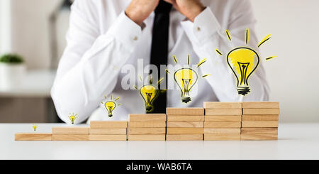 Businessman sitting at his desk with staircase made of wooden pegs and hand drawn light bulb above each step in a conceptual image.