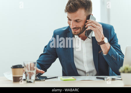 Young successful architect talking on smartphone and using digital tablet while working on the blueprints of a new project in the office. Stock Photo