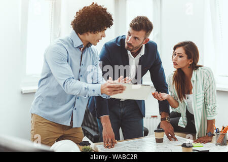 Team of three young architects using digital tablet and communicating while analyzing blueprints  of a new project in the office. Stock Photo