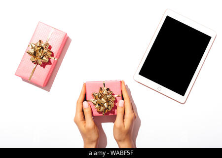 Flat lay woman's hands holding pink gift box next to digital tablet with blank screen on white background, top view. Stock Photo