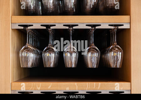 Closeup many upside down empty clear transparent crystal upturned wine glasses hanging in straight rows on brown wooden shelf, rack, showcase, stainle Stock Photo