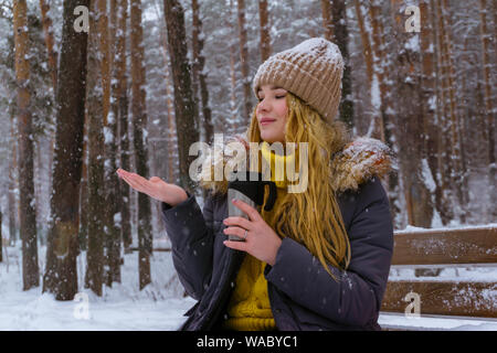 girl holding a thermomug and catches snowflakes with her hand while sitting on a bench in the winter park Stock Photo