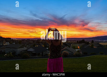 A woman takes a photo with a smartphone cell phone of a beautiful sunset over a suburb near Spokane, Washington. Stock Photo