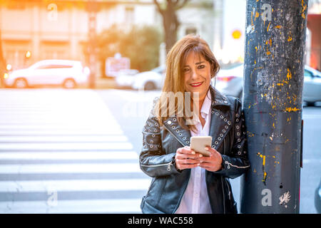 A smiling mature woman in a black leather jacket walking on the street and using her smartphone. Stock Photo