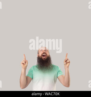 Portrait of surprised middle aged bald man with long beard in light t-shirt standing, looking with amazed face and pointing at up wall empty copyspace Stock Photo