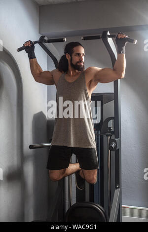 Full length portrait of confident young adult strong man with long curly hair working out in gym, hanging on horizontal bar with hands, doing pull ups Stock Photo