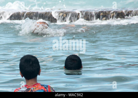 Three swimmers enjoying a swim in a tidal pool as the tide is coming in and the day is a warm sunny day. The two young ones are facing the elder one Stock Photo