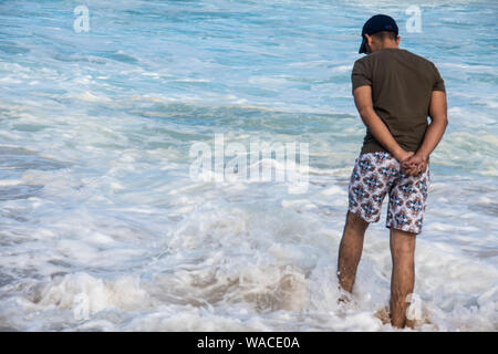 A man in casual beachwear pensively standing in ocean with hands behind his back whilst looking down in deep thought. The waves splashing on him Stock Photo