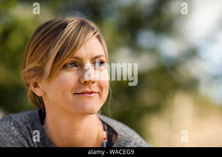 Smiling young woman looks thoughtfully into the distance as she sits outside. Stock Photo