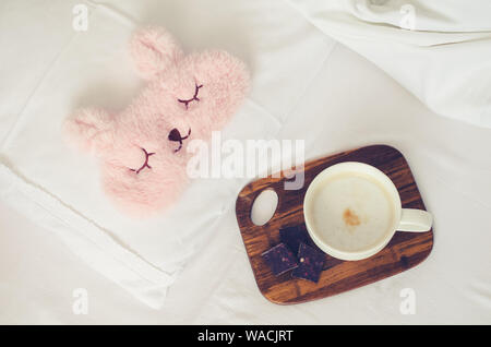 Cup of coffee with chocolate and cute pink sleep mask on the bed on cozy lazy sunday. White bedding sheet, blanket and pillows. Good morning concept. Stock Photo