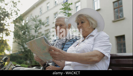 Senior two male and female tourists sitting on bench in town center with a map in hands and looking for the route while traveling in Lviv, Ukraine. Elegant woman in hat. Family vacation concept Stock Photo