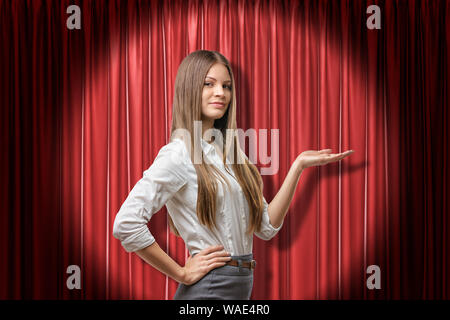 Young brunette business woman holding open palm up on red stage curtains background. Outfit ideas. Feelings and emotions. Gestures and body language Stock Photo