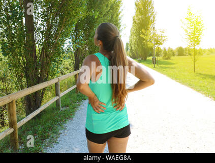 Back pain. Athletic running woman with back injury in sportswear rubbing touching lower back muscles standing outside in the park. Stock Photo