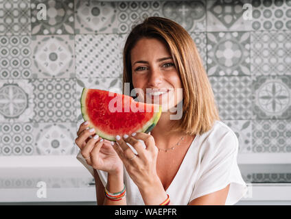Blonde woman with green eyes in modern kitchen part a watermelon to eat by hand enjoying it. Stock Photo
