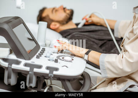 Men examining his thyroid with ultrasound sensor, lying in bathrobe on the couch at the medical office Stock Photo