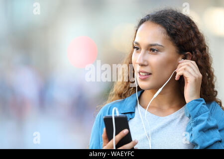 Mixed race woman holding smart phone putting earphones listening to music looking at side walking in the street Stock Photo