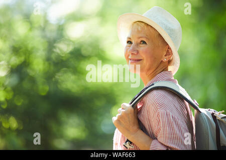 Smiling mature woman in hat with backpack behind her back standing outdoors and enjoying the good weather Stock Photo